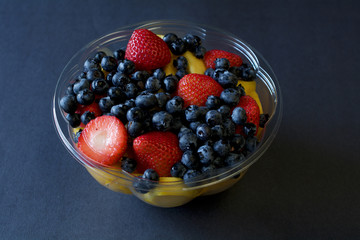 Mango, strawberries and blueberry fruit salad in plastic bowl against black background- healthy dessert concept 