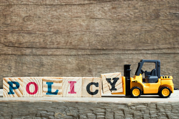 Yellow toy forklift hold letter block Y to complete word policy on wood background