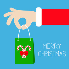 Merry Christmas. Santa Claus hand holding gift shopping paper bag with candy cane. Red costume fur. Giving present. Cute cartoon kawaii funny father character. Blue background. Greeting card. Flat