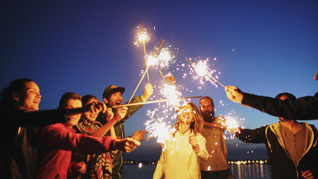 Group Of Young Friends Having A Beach Party. Friends Dancing And Celebrating With Sparklers In Twilight Sunset