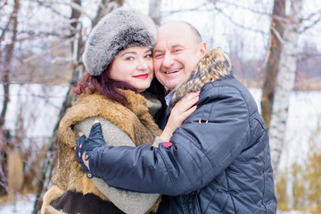 Obraz premium Portrait of happy stylish couple hugging in snowy park. Happy mature people in winter clothes embracing on nature background. Happy senior couple walking in winter outdoors 