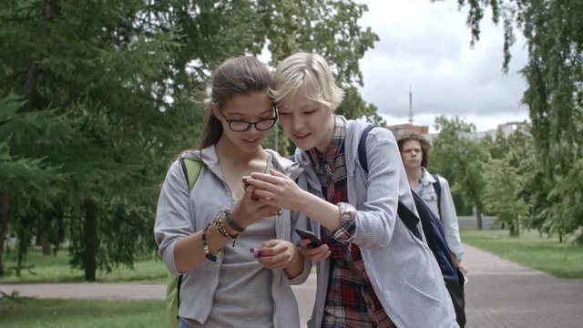 Two young girls walking together in park at summer day, using smartphones and discussing social media posts; teen boy walking behind them with digital tablet