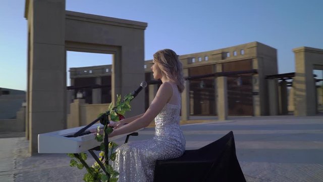 Elegant young blonde woman sings, plays romantic music on white digital piano in big arena or amphitheater. Girl in shiny evening dress performs on stage. Dubai Bab Al Shams Arena