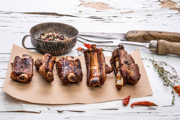 Grilled pork ribs on piece of brown paper with herbs, spices and carving fork on white wooden table with cracked paint.