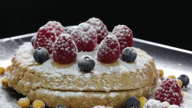 Cake with raspberries, blueberries, sea buckthorn sprinkled with powdered sugar on a black plate rotates