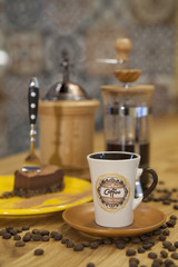 A cup of coffee and a piece of cake placed on a table with a coffee maker in the background