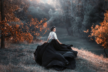 girl in black and white vintage dress is running looking around. Silk fabric skirt train waving fly in wind motion. Artistic Photography. Medieval fantasy woman walk in autumn nature, tree orange leaf