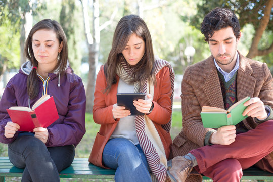 One Woman Sitting On A Bench In A Park Reading An E-Book With Two People Reading Paper Books