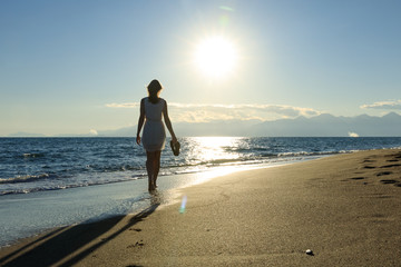 Young, sexy woman is walking at a beautiful sunset beach. holding her flip flops in her hand. sexy short dress skirt. wet feet legs. sundown mountains