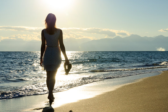 Junge, sexy Frau läuft flaniert am Strand Meer entlang. In der Hand hält sie Flip Flops. Träumend Selfie mit Handy, Welliges Meer und oranger Sonnenuntergang in den Bergen