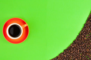 Lot of roasted coffee beans scattered on green paper beside full ceramic cup of black coffee on orange saucer