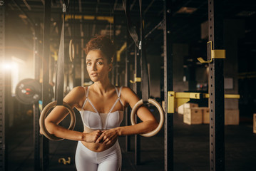 Muscle-up exercise athletic woman doing intense workout at the gym on gymnastic rings. Fit young woman holding gymnast rings at the gym looking away. Young caucasian woman exercising at gym.