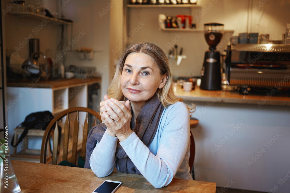 Wall mural Beautiful middle aged female wearing scarf sitting at coffee shop, holding mug of hot drink with both hands, warming up after morning walk outdoors on cold autumn day, looking and smiling at camera