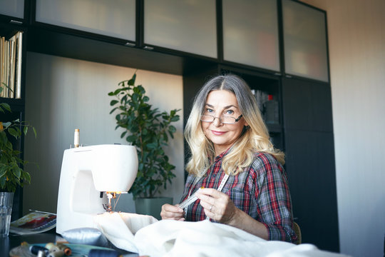 Indoor view of attractive blonde woman pensioner making her living by sewing, repairing and altering clothing items, working from home, sitting in front of sewing machine, stitching white fabric