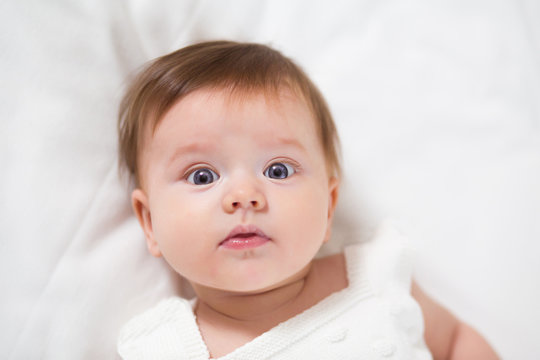 Top view and close-up portrait of cute adorable baby girl wearing white body in bedroom looking at camera. Newborn child relaxing and smiling in the bed in children nursery. Family morning at home