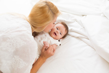 Happy loving family concept. Beautiful mother playing with her baby girl in the bedroom. They smiling and hugging together on white bed linens