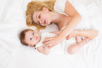 Obraz na płótnie Canvas Portrait of young mother with blond hair with her sweet 3 month old baby girl in white wear having fun in the bedroom at morning, loving happy family concept, top view