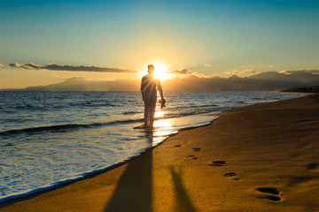 Junger, sexy Mann läuft, spaziert barfuß am Strand durchs Wasser in den Sonnenuntergang. Hält dabei seine Flip Flops in der Hand. Berglandschaft am Horizont blaue Farben