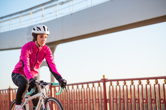 Young Woman in Pink Jacket Riding Road Bicycle on the Bridge Bike Line in the Cold Sunny Autumn Day. Healthy Lifestyle.