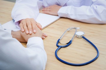 friendly female doctor holding patient hand for reassuring with clipboard and stethoscope on desk in hospital, people, age, health, care and support concept, selective focus