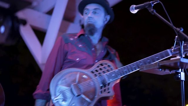 Closeup of guitarist finishing up a song on a resonator at a live performance outside at night.