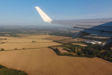  Sun-shining passenger plane wing against a blue sky and yellow-brown green fields on the ground