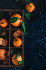 Tangerines in a wooden case on a dark background. An assortment of clementines with green leaves. Dark food photography with vibrant orange fruit and copy space.