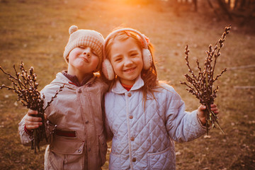 Little boy and girl stand with willow branches outside in the rays of evening sun