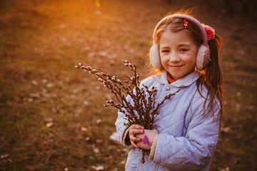 Charming little girl in blue jacket stands on the fallen leaves outside with willow branches