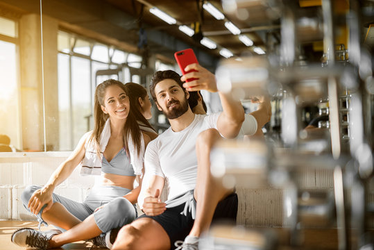 Young attractive smiling active fitness couple sitting on the floor of the gym and taking a selfie, leaning on the mirror with a towel.