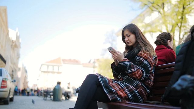 A girl is sitting on a bench and typing messages using smartphone.Outdoor,spring