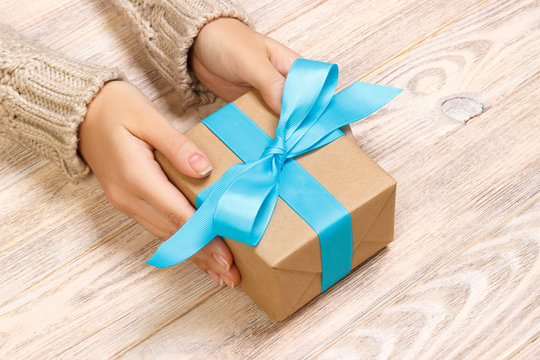 Woman's hands give wrapped valentine holiday handmade present in craft paper with blue ribbon. Present box, decoration of gift on black wooden table, POV with copy space