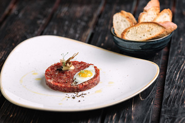 Beef tartare with fried quail egg served with bread on dark wood background