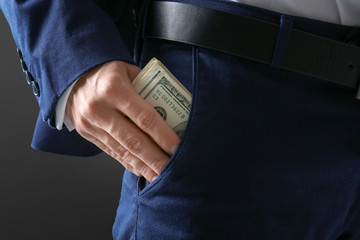 Man in formal suit putting money in pocket on dark background, closeup - obrazy, fototapety, plakaty
