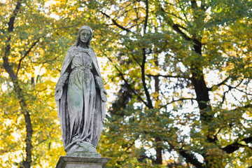 Fototapeta premium Statue of Mary Mother of God, at autumn tree backgroung, copy space. Religious symbol.
