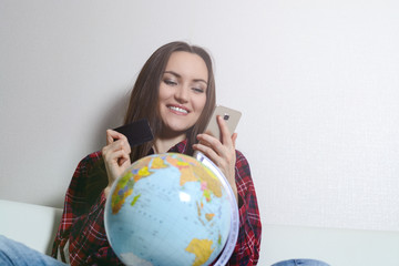 Happy woman with credit card and phone in hand, smiling sitting on sofa with globe. The joy of buying tickets for travel