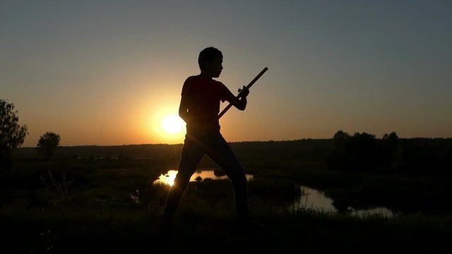 Boy Strikes Kung-Fu Blows With a Stick on a Lake Bank at Sunset