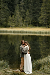 Smiling bride and groom stand and embrace near forest lake.