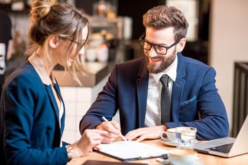 Business couple dressed stricly in the suits signing a contract sitting with coffee and laptop at the cafe or restaurant