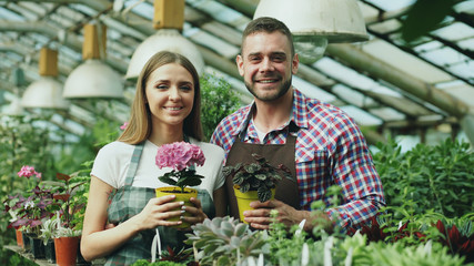 Happy young couple smiling in greenhouse. Attractive woman and man florists in apron work in garden looking into camera