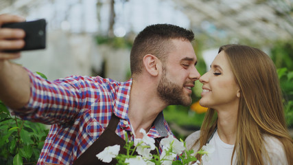 Cheerful loving couple gardeners taking selfie picture on smartphone camera and kissing while working in greenhouse