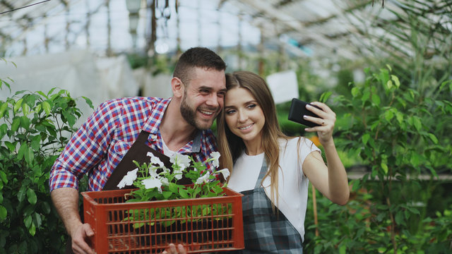 Cheerful loving couple gardeners taking selfie picture on smartphone camera while working in greenhouse