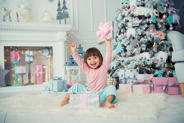 Positive cheerful baby girl sitting with Christmas gift near Christmas tree. Happy New Year