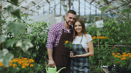 Happy young couple smiling in greenhouse. Attractive woman and man florists in apron work in garden...