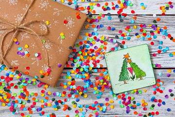 Obraz premium Present box with a white painted pattern on white wooden background with multicolored confetti and a card with two loving firs. Flat lay style. Christmas New Year Celebration Decorations