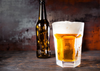 Empty beer bottle next to glass with a light beer and a head of foam on old dark desk