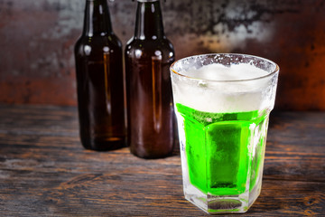 Two beer bottles next to glass with a green beer and a head of foam on old dark desk