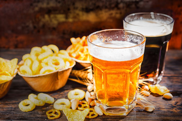 Two glasses with light and dark beer near plates with chips and scattered snacks on dark wooden desk