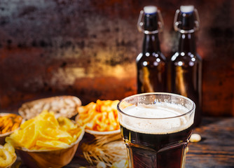 Plates with snacks near two bottles and a glass of dark beer, wheat, scattered nuts and pretzels on dark wooden desk