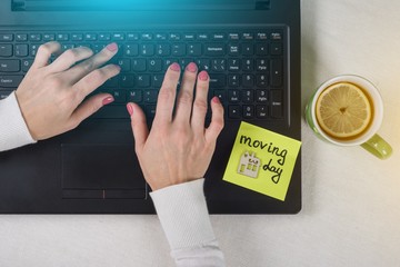 The concept of new housing, moving. A note of text Moving Day written on the paper sticker. Laptop, computer woman's hands background.